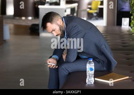 Geschäftsmann, der kurze Pause macht, sitzt in der Lobby des Büros mit Smartphone Stockfoto