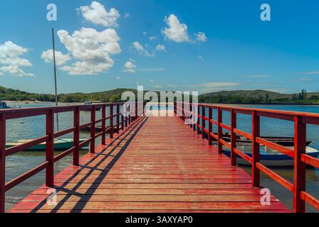 Ein pulsierender roter Holzpier erstreckt sich in die ruhige Lagune unter einem hellblauen Himmel, mit kleinen Booten entlang und grünen Hügeln am Horizont. Stockfoto
