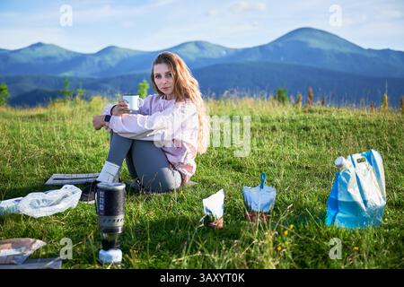 Die Touristin sitzt auf einem grasbewachsenen Hügel und hält den Becher. Reisender mit langen blonden Haaren. In der Nähe tragbarer Campingkocher, Essensbeutel, blaue Tasche. Die malerische Bergkulisse sorgt für eine ruhige Atmosphäre im Freien. Stockfoto
