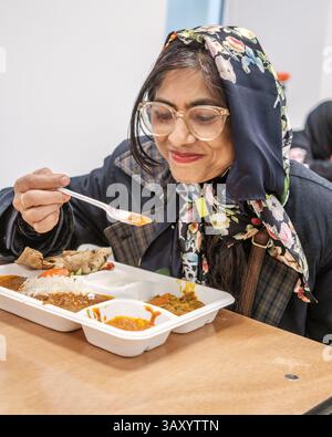 Eine Indianerin mit Brille und geblümtem Kopftuch sitzt an einem Tisch und isst von einem Tablett mit traditionellem indischem Essen. Stockfoto