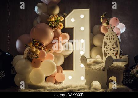 Bezaubernde Fotozone für den ersten Geburtstag des Babys: Rosafarbene Ballons, beleuchtete Zahl „1“, weiche weiße Basis und gemütliche warme Töne Stockfoto