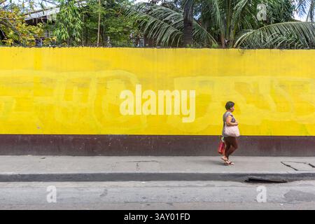 Eine Frau läuft an einer breiten gelben Mauer in Manila, Philippinen, vorbei und fügt den lebendigen Farben und Texturen der städtischen Straßenlandschaft ein menschliches Element hinzu Stockfoto