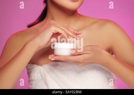 Frau, die Hautpflegecreme in einem sanft beleuchteten Raum mit violettem Hintergrund aufträgt Stockfoto
