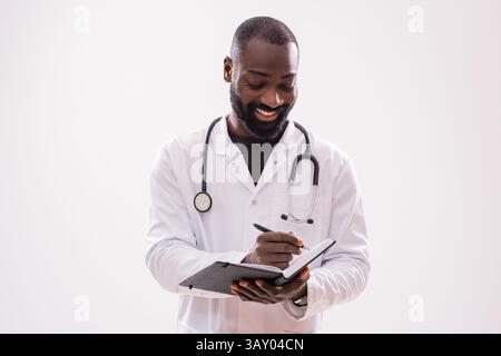 Porträt eines fröhlichen schwarzen männlichen Sanitäters in medizinischer Uniform, der Notizen im persönlichen Organisator schreibt. Studio-Aufnahme auf grauem Hintergrund. Stockfoto