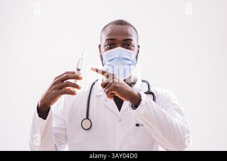 Porträt eines afrikanischen Arztes in medizinischer Uniform mit Schutzmaske auf Spritze auf grauem Hintergrund. Stockfoto