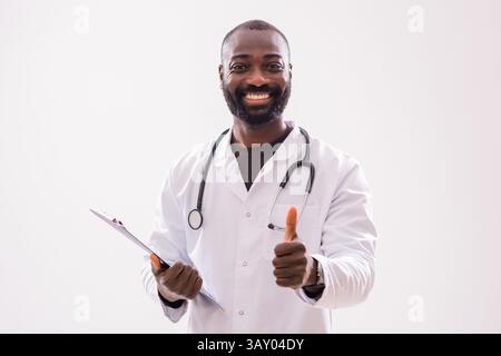 Porträt eines fröhlichen afroamerikanischen Arztes in medizinischer Uniform, der Clipboard hält und Daumen auf grauem Hintergrund zeigt. Stockfoto