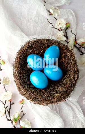 Nest mit blau bemalten Ostereiern, zarten weißen Blüten und weichem rosa Stoff auf weißem Hintergrund Stockfoto