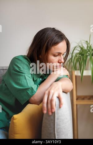 Traurige und müde Frau, die zu Hause auf dem Sofa sitzt und nach unten blickt Stockfoto