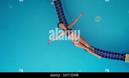Luftaufnahmen eines jungen Touristen, der einen schwarzen Bikini trägt und in einem blauen Schwimmbadwasser mit offenen Armen in einem tropischen Resort schwimmt Stockfoto