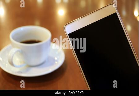 Nahaufnahme eines Mobiltelefons mit leerem Bildschirm und unscharfem schwarzem Kaffee im Hintergrund Stockfoto