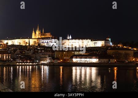 Fesselnder nächtlicher Blick auf die Prager Burg, die unter den Lichtern leuchtet, mit historischer Architektur, die sich am Himmel befindet. Der beschauliche Fluss reflektiert die Stockfoto