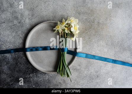 Blick von oben auf einen Strauß weißer Narzissen, ordentlich mit einem dunkelblauen Band gebunden, auf einer runden grauen Platte vor einem strukturierten grauen Hintergrund. Th Stockfoto