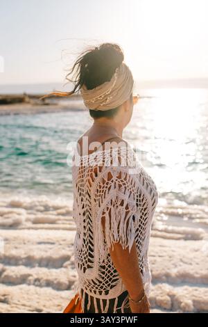Seitenansicht einer nicht erkennbaren Frau mit Kopftuch, die in Richtung des sonnendurchfluteten Horizonts über dem Toten Meer in Jordanien blickt und somit Gelassenheit und Fernweh symbolisiert. Stockfoto