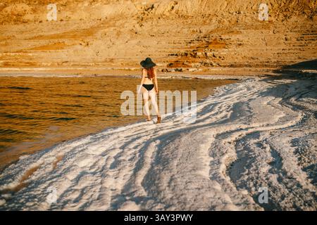 Rückansicht einer nicht erkennbaren Frau in Bikini und breiter Krempe, die entlang der Salzformationen am Toten Meer in Jordanien spaziert. Stockfoto