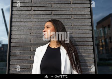 Eine selbstbewusste Geschäftsfrau in weißem Blazer und schwarzem Oberteil steht vor einer grauen Ziegelwand. Das Bild spiegelt einen professionellen und modernen Lebensstil wider Stockfoto