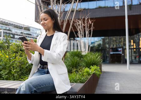 Hispanische Frau in einem weißen Blazer und Jeans benutzt ein Smartphone vor einem modernen Büro. Sie vertritt das berufliche Vertrauen in ein städtisches Unternehmen Stockfoto