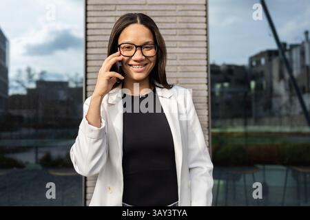 Eine selbstbewusste hispanische Frau in einem weißen Blazer und einer Brille lächelt, während sie auf ihrem Smartphone spricht. Sie steht in einer modernen urbanen Umgebung und verkörpert einen Profi Stockfoto