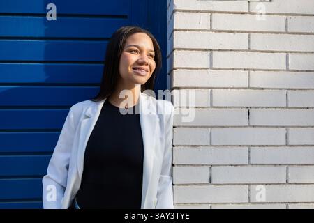 Selbstbewusste Frau mit Zahnspangen in einem weißen Blazer, die vor einer modernen blau-weißen Wand steht. Sonnendurchflutetes Gesicht, das Positivität und Professionalität ausdrückt Stockfoto