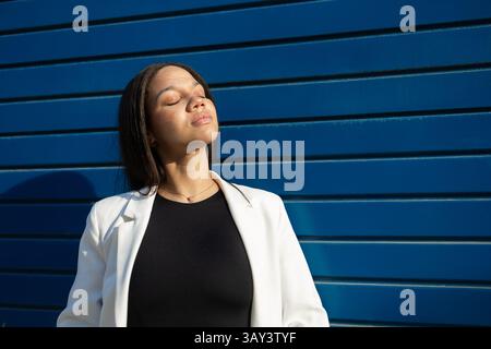 Eine selbstbewusste Frau in einem weißen Blazer sonnt sich im Sonnenlicht an einer blauen Wand. Ihre geschlossenen Augen und ihr heiterer Ausdruck verkörpern ein Gleichgewicht zwischen Geschäft und Rela Stockfoto