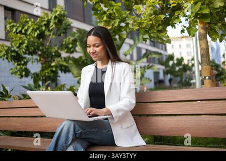 Eine hispanische Geschäftsfrau in einem weißen Blazer arbeitet an ihrem Laptop, während sie auf einer Bank in einer grünen städtischen Umgebung sitzt. Sie scheint fokussiert und Professi zu sein Stockfoto