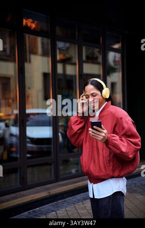 Ein Mann, der gelbe Kopfhörer trägt, ein Smartphone hält und lächelt, während er draußen Musik genießt. Sie tragen eine rote Jacke, stehen an einem Glasgebäude Stockfoto