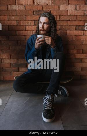 Ein Mann mit langen Haaren und Bart sitzt auf dem Boden vor einer Ziegelwand und hält eine Tasse Kaffee. Er scheint tief in Gedanken zu sein und trägt legere Kleidung Stockfoto