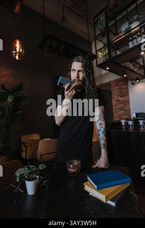 Bärtiger Mann mit Tattoos, der ein Smartphone in einem stilvollen Café benutzt. Warme Beleuchtung und Pflanzen schaffen eine gemütliche Atmosphäre, mit einem Tisch, auf dem eine Pflanze gehalten wird, Stockfoto