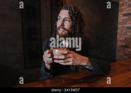 Ein Mann mit langen Haaren und Bart sitzt an einem Holztisch und hält eine Tasse Kaffee mit Latte Art. Die Umgebung ist ein gemütliches Café mit einem warmen und entspannten A Stockfoto