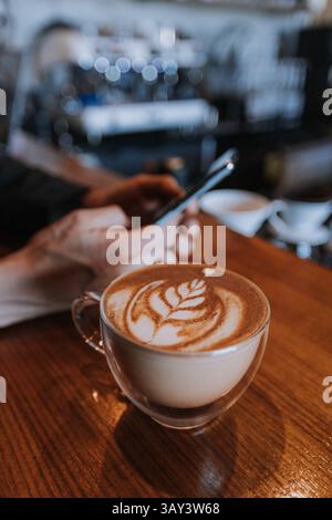 Ein Mann benutzt ein Smartphone in einem Café und konzentriert sich auf einen wunderschön gefertigten Latte mit komplizierter Kunst. Das warme Ambiente verstärkt die gemütliche, entspannte Atmosphäre Stockfoto