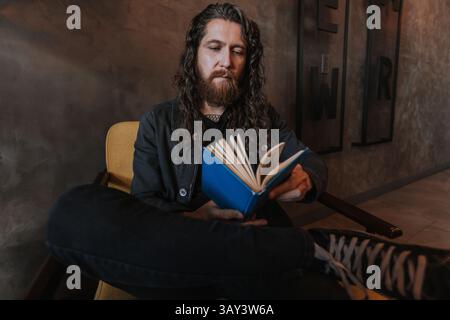 Ein Mann mit langen Haaren und Bart liest ein blaues Buch, während er in einem gemütlichen Stuhl eines Cafés sitzt, was eine entspannte und intellektuelle Atmosphäre schafft, perfekt für ihn Stockfoto