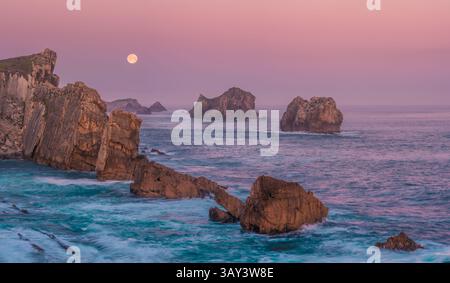 Ein ruhiger Blick auf zerklüftete Seestapel und Klippen im sanften Morgenlicht am Strand von Arnia, Costa Quebrada, mit Vollmond über der ruhigen A Stockfoto