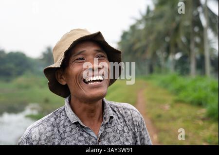 Ein fröhlicher indonesischer Bauer steht in einem üppigen Reisfeld mit Hut und gemustertem Hemd. Die ruhige Landschaft mit Grün und Palmen, Symb Stockfoto