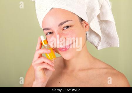 Eine Frau mit einem Handtuch um den Kopf hält eine kleine Flasche Gesichtsserum. Ihr strahlender Teint und ihr sanftes Lächeln spiegeln eine pflegende Hautpflege wider Stockfoto