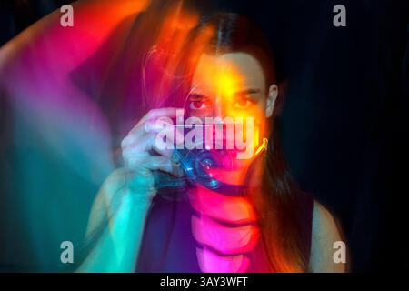 Ein kreatives Porträt einer Frau, die eine Kamera hält, aufgenommen mit Langzeitbelichtung. Farbenfrohe Lichtspuren umgeben die Frau und schaffen eine dynamische und künstlerische Atmosphäre Stockfoto