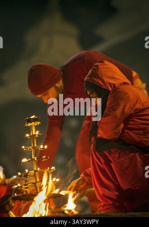 Hindu-Priester, der abends aarti (rituelle Anbetung mit Feuer) in einem Tempel oder am Ufer vorführt, in roten Gewändern gekleidet und von Öllampen umgeben Stockfoto