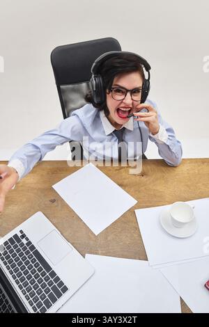 Junge Frau in formellem Outfit, die während der Arbeit am Laptop in das Headset schreit und Frustration und Wut in chaotischen Büroumgebungen zeigt. Stockfoto