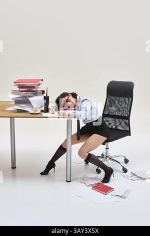 Junge Frau, die Kopf auf Dokumenten ruht, sich mit verschlafenem Ausdruck auf den Schreibtisch lehnt, in unordentlichem Büro-Setup vor weißem Studio-Hintergrund. Stockfoto