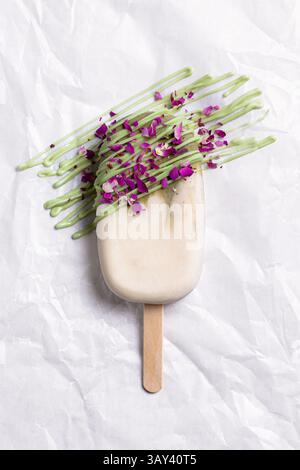 Ein Eis mit Vanille- und Matcha-Geschmack, das mit lebendigen essbaren Blumen und einem grünen Niesel geschmückt ist und auf einem zerknitterten weißen Papier präsentiert wird Stockfoto