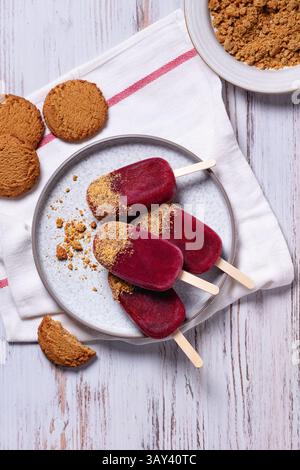 Blick von oben auf lebendige Himbeeren und Kekse gewürzte Eispops, die auf einem Metalltablett serviert werden, umgeben von zerquetschten Keksen auf einem rustikalen Holztisch. Stockfoto