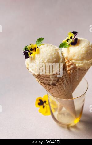 Zwei Kugeln Vanilleeis, serviert in Waffelkegeln, geschmückt mit kleinen essbaren Stiefmütterchenblüten und Minzblättern, in einem Glas. Stockfoto