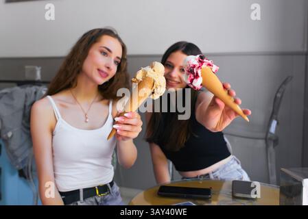 Zwei junge Frauen genießen gerne Eiskegel in einem Café. Sie lächeln, während sie verschiedene aromatisierte Kegel halten, um einen Moment der Freude und der Pommes festzuhalten Stockfoto