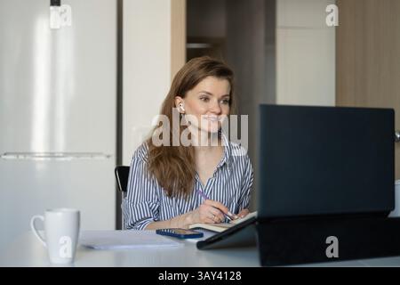Eine Frau sitzt an einem Tisch und arbeitet von zu Hause aus. Sie schreibt in einem Notizbuch, während sie sich auf ihren Laptop-Bildschirm konzentriert, mit einem Becher und einem Telefon in der Nähe. Stockfoto