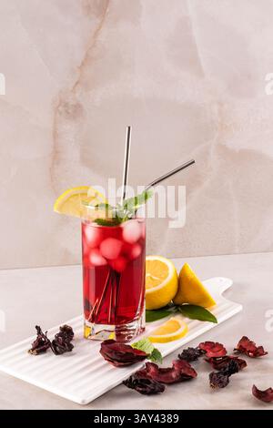 Ein lebendiges Glas Hibiskustee mit Eis, garniert mit Zitronenscheiben und Minze. Es sitzt auf einer hellen Marmoroberfläche und schafft eine erfrischende und einladende Atmosphäre Stockfoto