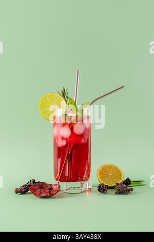 Ein lebendiges Glas Hibiskus-Eistee mit Limettenscheiben, Minzblättern und Eiswürfeln vor pastellgrünem Hintergrund bietet ein erfrischendes Erlebnis Stockfoto