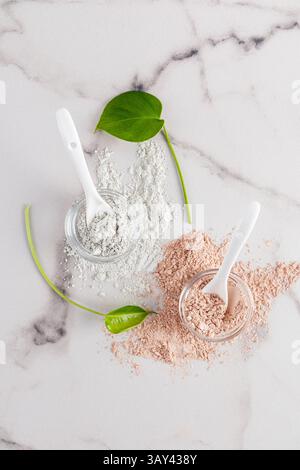 Draufsicht auf zwei Arten kosmetischer Tonpulver in klaren Gläsern mit Löffeln und grünen Blättern auf weißem Marmorhintergrund. Stockfoto