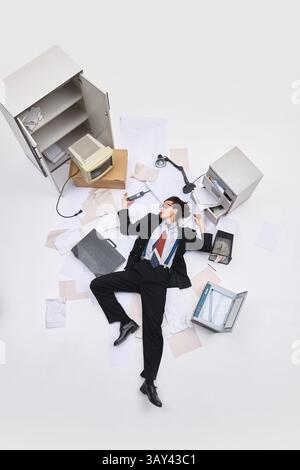 Geschäftsmann in lockerer Krawatte, der sich auf dem Boden mit Telefon in der Hand ausbreitete und Papier um den Körper in einem chaotischen weißen Büro verstreute. Stockfoto