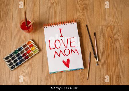 Eine herzliche „Ich LIEBE MAMA“-Nachricht ist rot auf einem spiralförmigen Notizbuch gemalt, umgeben von Pinseln, Aquarellpalette und einem Malbecher, wodurch ein warmes A entsteht Stockfoto