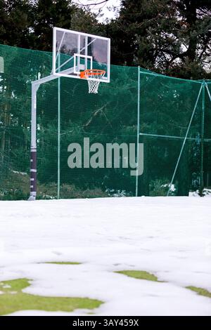 Ein Basketballkorb steht am Rande eines schneebedeckten Außenplatzes mit Waldkulisse. Die Szene fängt die Ruhe eines Wintertages ein Stockfoto