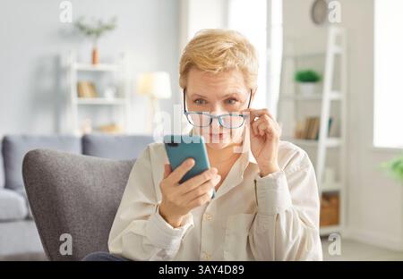 Reife Frau mit schockiertem Ausdruck und leicht abgesenkter Brille schaut auf ihren Handy-Bildschirm. Stockfoto