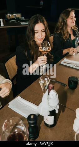 Eine Frau sitzt an einem Tisch mit einem Glas Wein vor sich. Sie lächelt und genießt ihren Drink. Es gibt noch andere Leute im Backgr Stockfoto